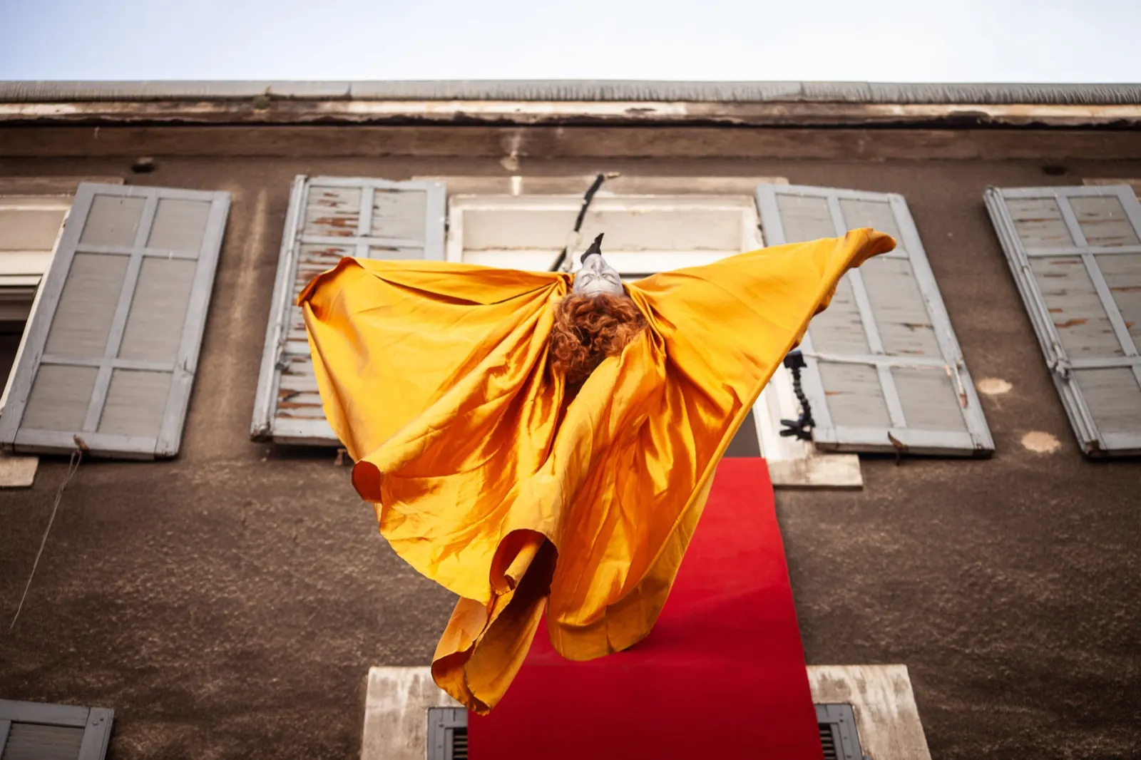 A dancer in billowing fabric hangs on a building façade above a red carpet